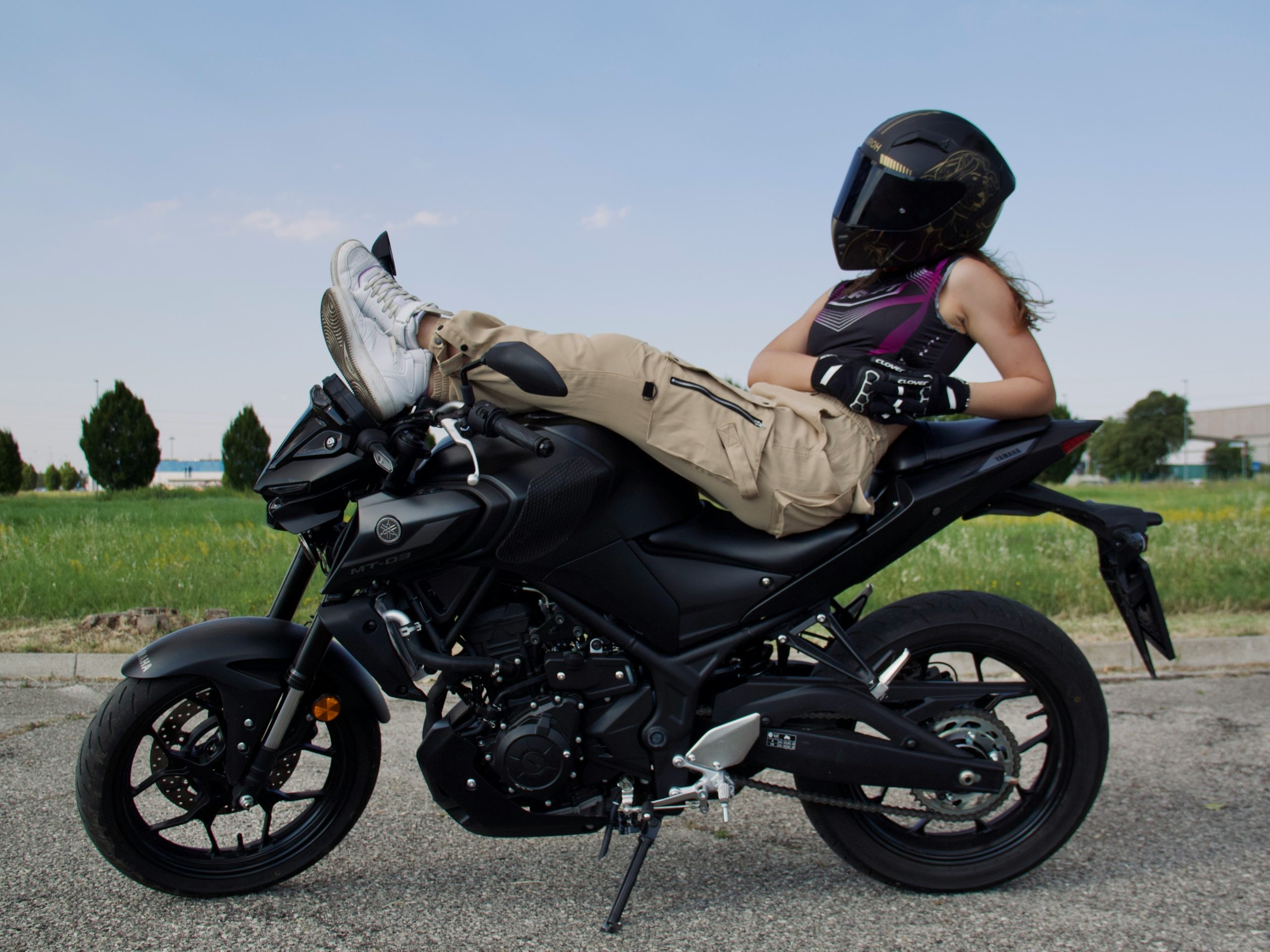Fille casquée et gantée assise sur sa moto, accoudée au siège passager, pieds sur le guidon